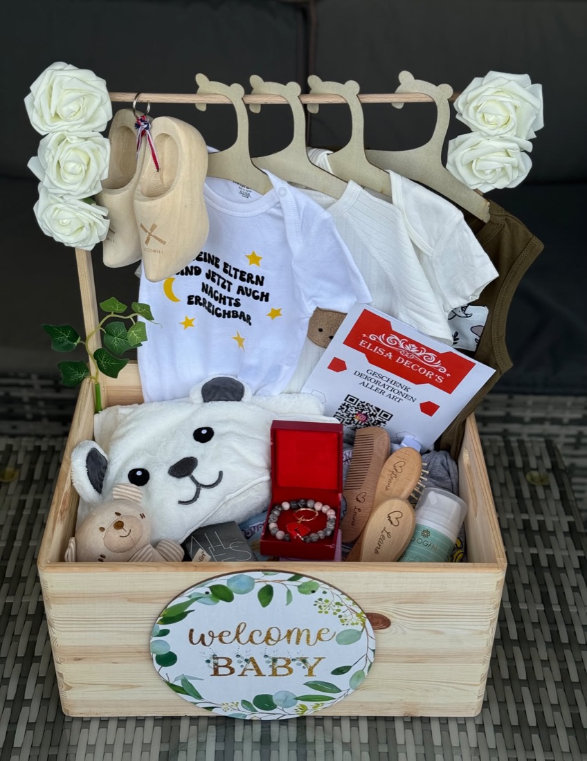 Holzkiste mit Babykleidern, Plüschtier und Schild Welcome Baby